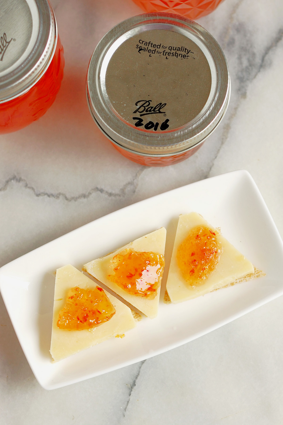 a jar of ghost pepper jelly. A white plate with crackers with cheese and jelly.