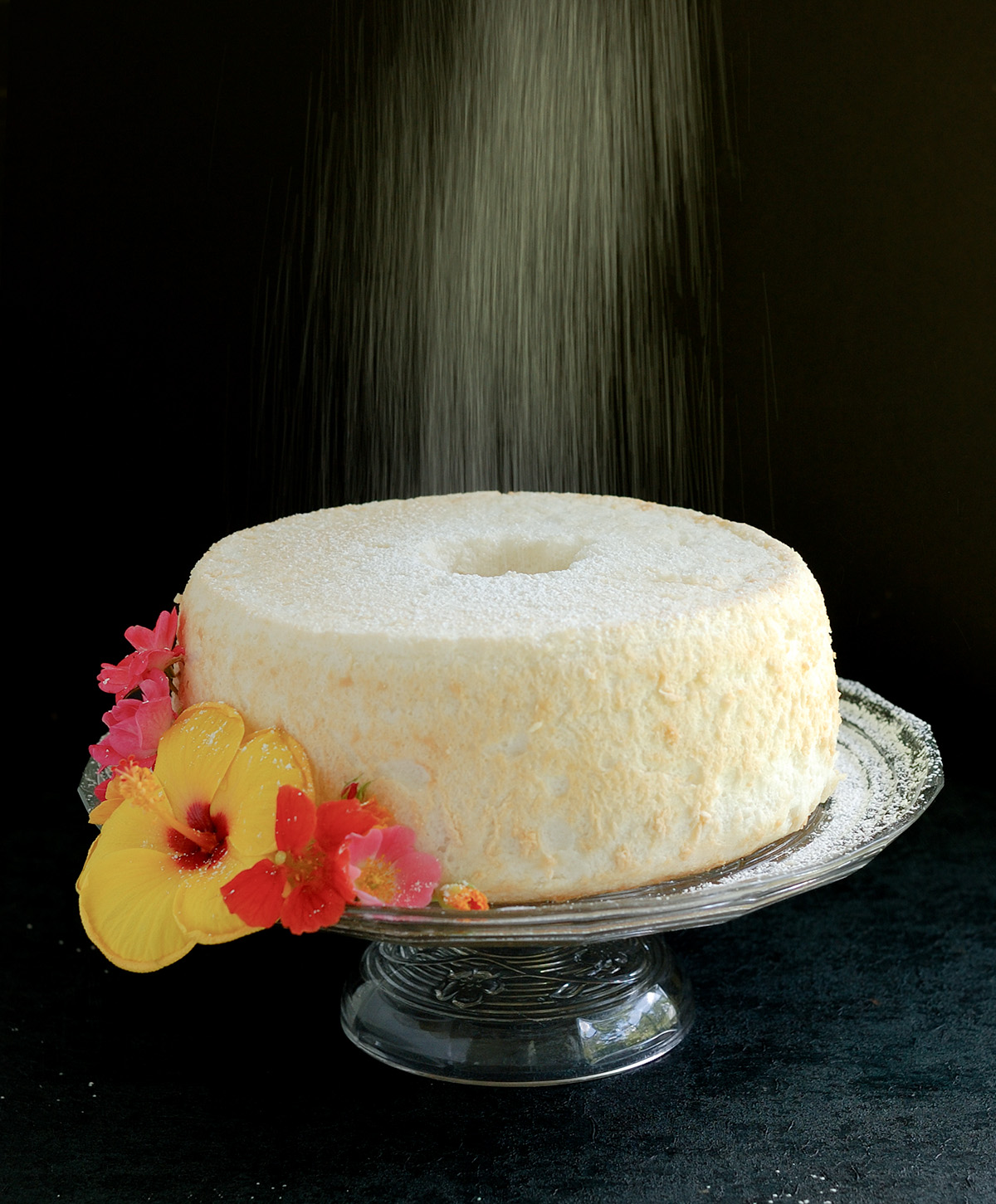 an angel food cake on a glass cake stand with sprinkled sugar and flowers.