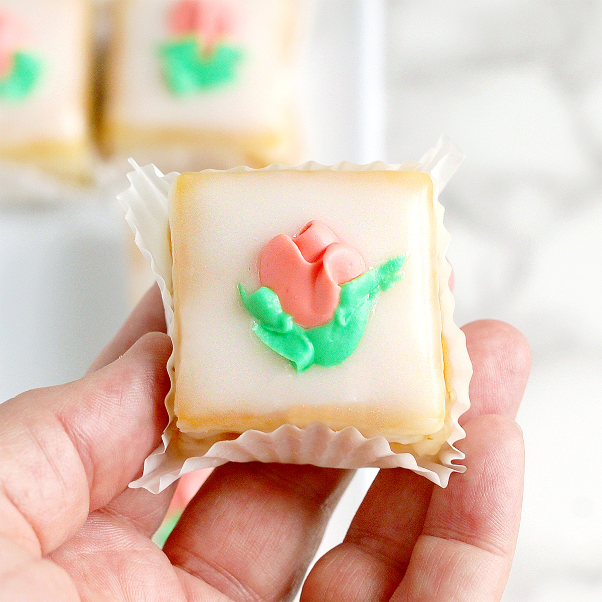 a hand holding a petit four with a pink flower.