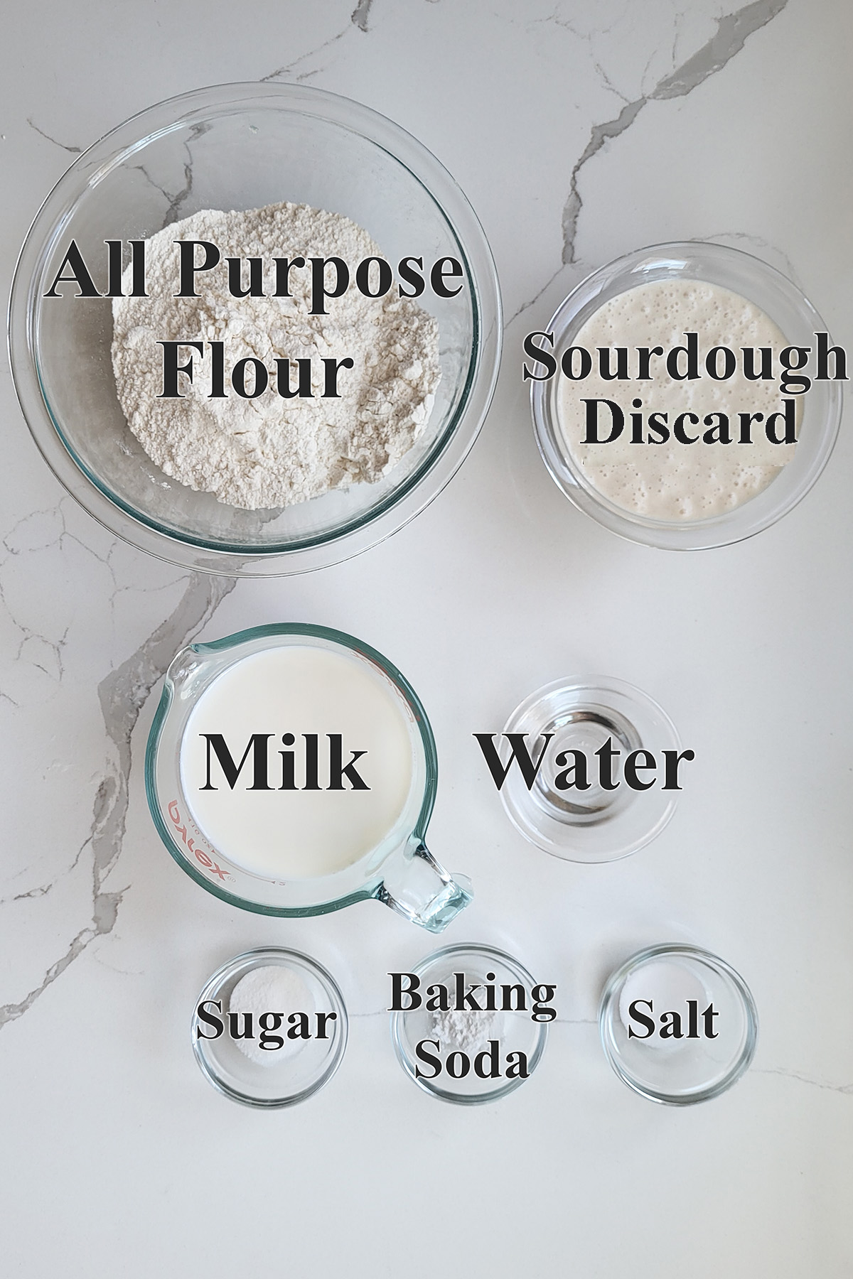 ingredients for sourdough crumpets in glass bowls on marble surface.