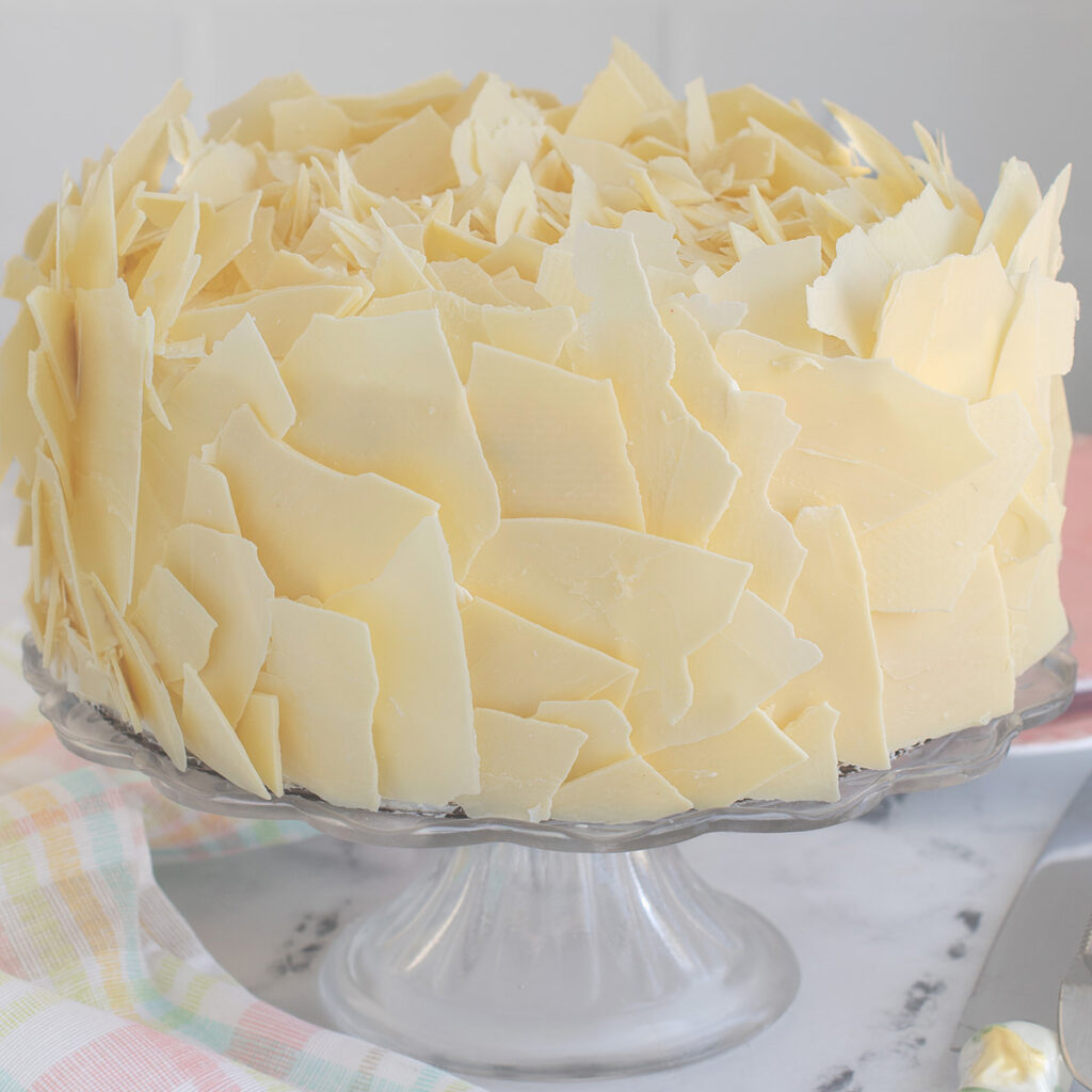 a white chocolate cake covered in white chocolate flakes on a glass cake stand.